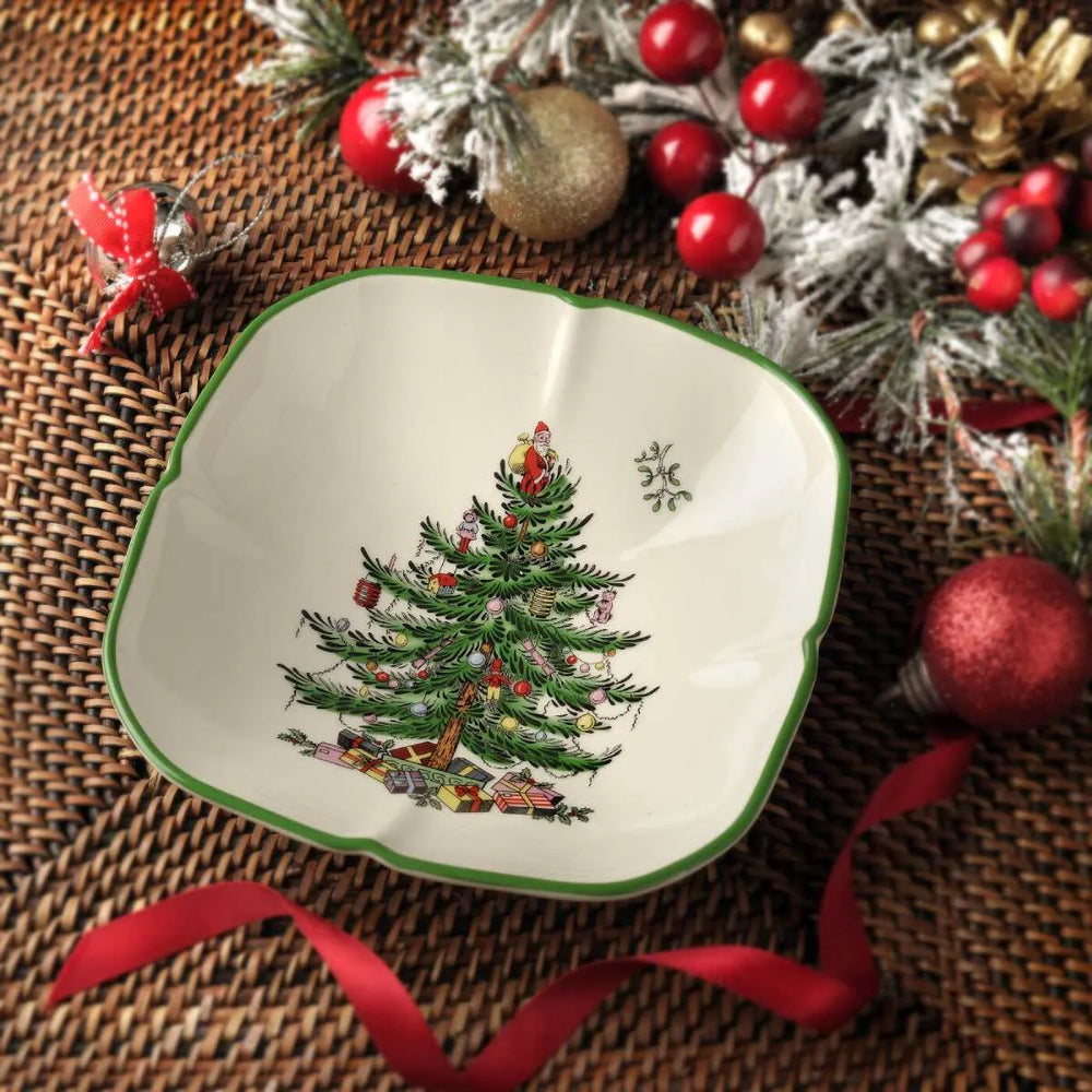 A square ceramic bowl with scalloped edges, decorated with a green Christmas tree illustration and a green rim, styled on a woven brown mat with holiday ornaments and ribbons.