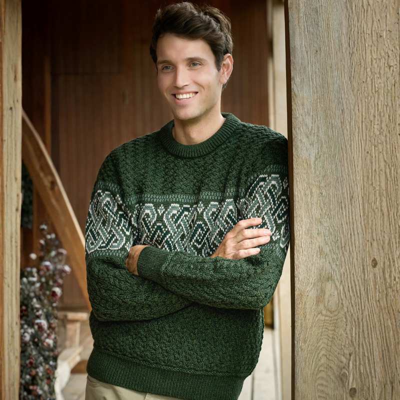 Man wearing a green sweater with a pattern, standing indoors against wooden walls.