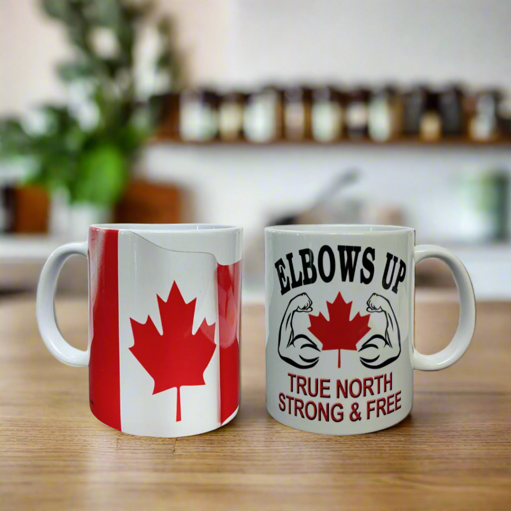 Two white mugs with red and black text, one featuring a red maple leaf and the other with a phrase 'ELBOWS UP TRUE NORTH STRONG & FREE' on a kitchen counter.