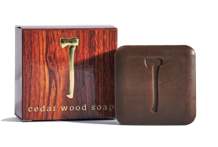 A bar of cedar wood soap in a dark brown leather-like square block with an embossed axe symbol, shown next to its matching reddish-brown wood-grain box with a gold axe emblem and the text 'cedar wood soap'.