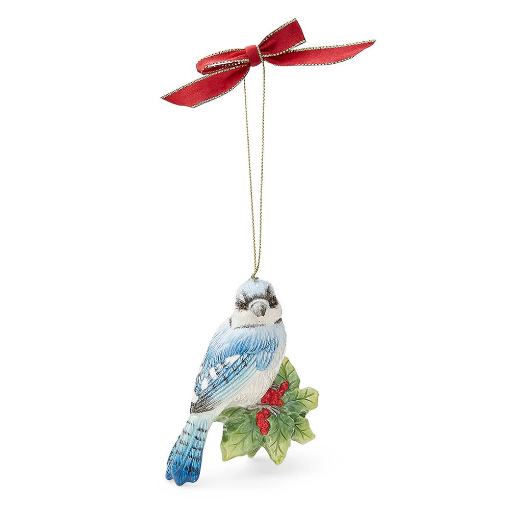 Blue jay ornament with bright blue feathers, perched on holly leaves with red berries. It hangs from a golden string topped with a red ribbon bow.