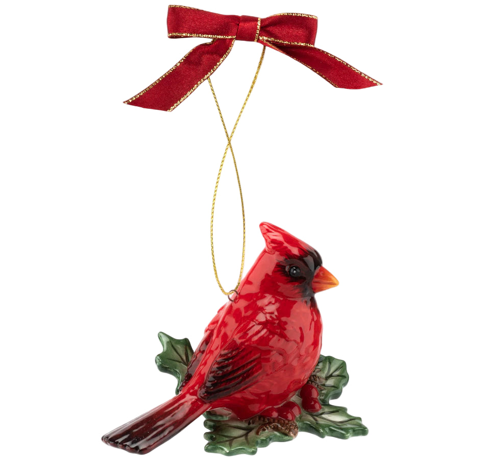 Red cardinal ornament with glossy finish, perched on green holly leaves. It hangs from a gold string, adorned with a red bow, conveying festivity.