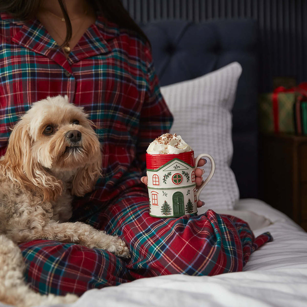A person in red plaid pajamas sits on a bed, holding a festive mug topped with whipped cream. A small, fluffy dog rests beside them. Cozy and warm ambiance.