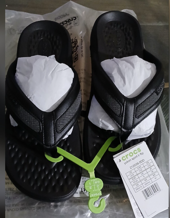 A pair of navy Crocs flip-flop sandals in their original plastic packaging, with white tissue paper inside the footbeds and a green plastic tag attached. A Crocs retail tag is visible on the right side.