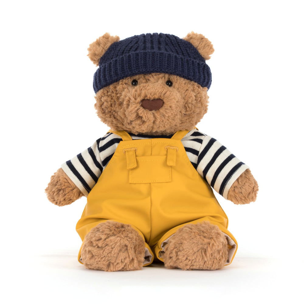 Teddy bear wearing a navy blue hat, striped shirt, and yellow overalls on a white background