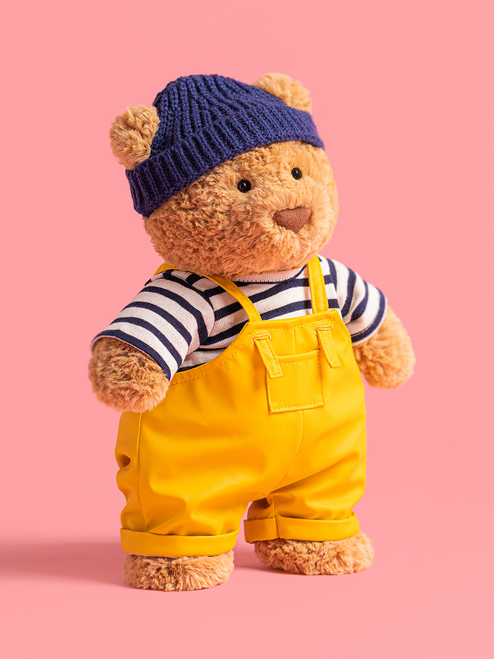 Teddy bear wearing a blue knitted hat, striped shirt, and yellow overalls on a pink background