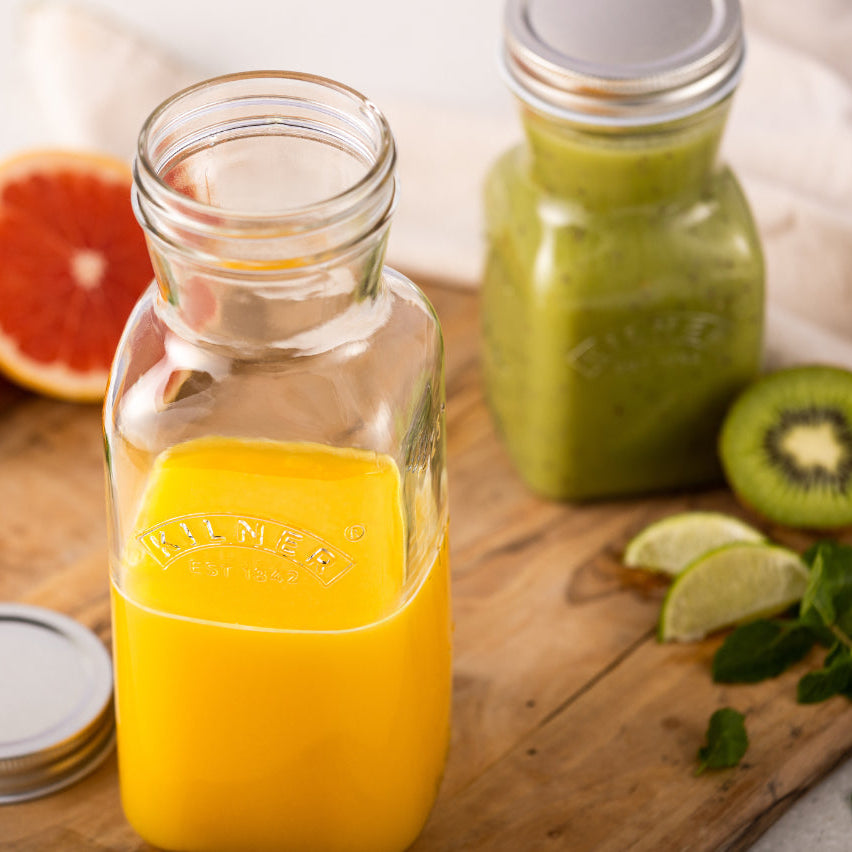 Two glass jars with juice on a wooden surface, surrounded by fruits and herbs.
