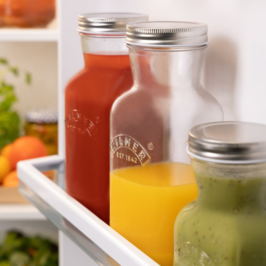Three glass jars with silver lids containing different colored liquids on a shelf with fruits and vegetables in the background.
