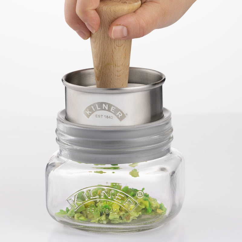 Clear glass jar with wooden and metal component in a white background mashing avocado 