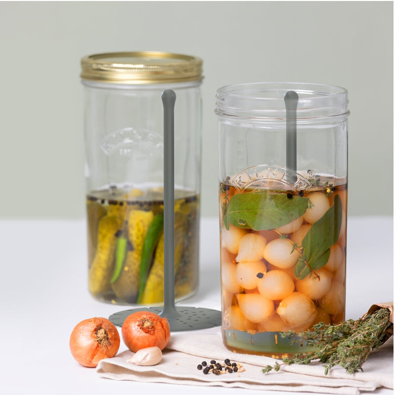 Two jars filled with pickles and garlic surrounded by herbs and onions