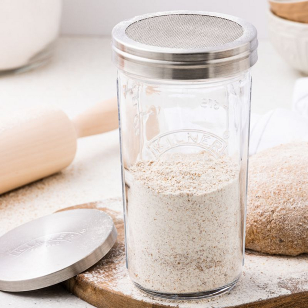 A clear glass sifter jar with a stainless steel lid and a mesh sifter compartment, surrounded by bread flour