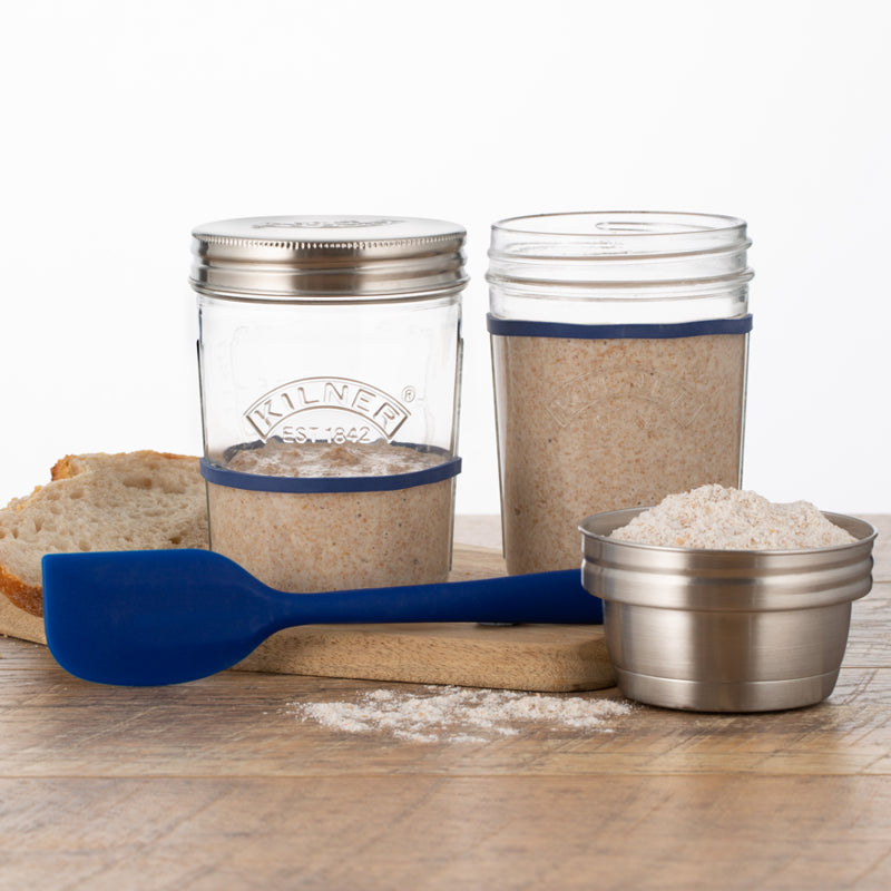Two glass jars with metal lids, a blue spatula, and a small metal container on a wooden surface.