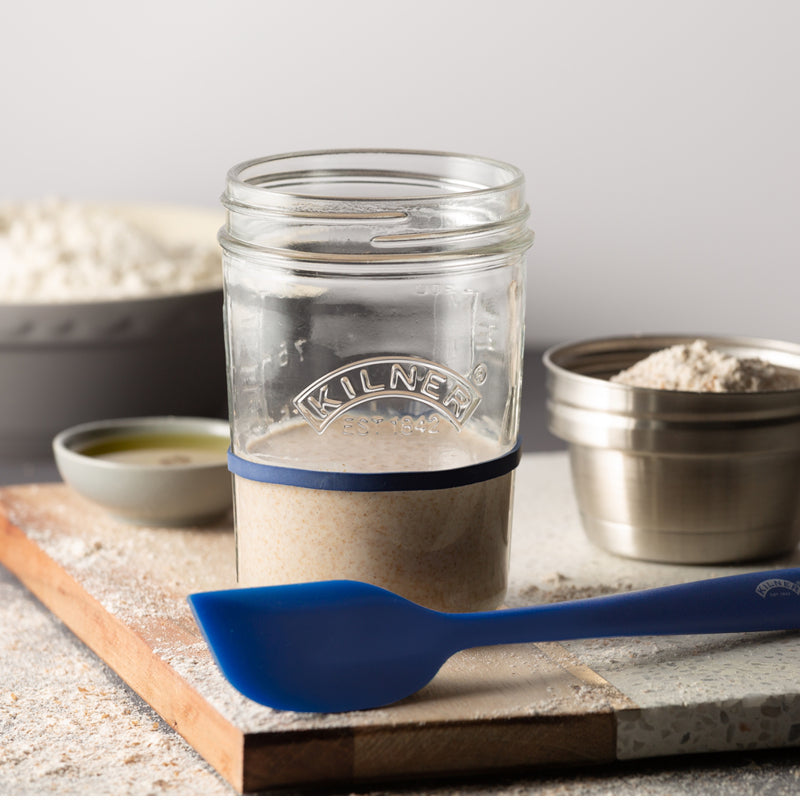 Clear glass jar with a blue band labeled 'Kilner' on a wooden surface with flour and a blue spoon.