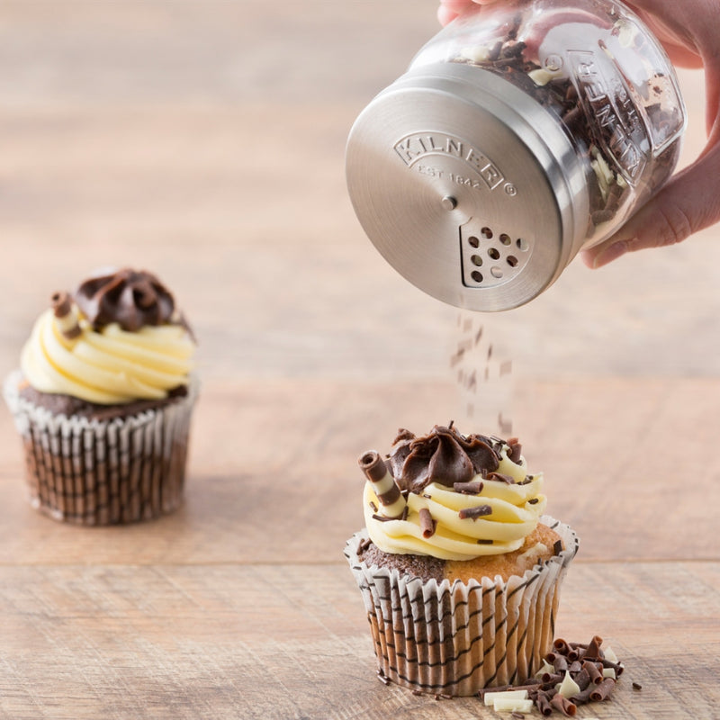 Cupcake being sprinkled with chocolate shavings from a Kilner jar on a wooden surface.