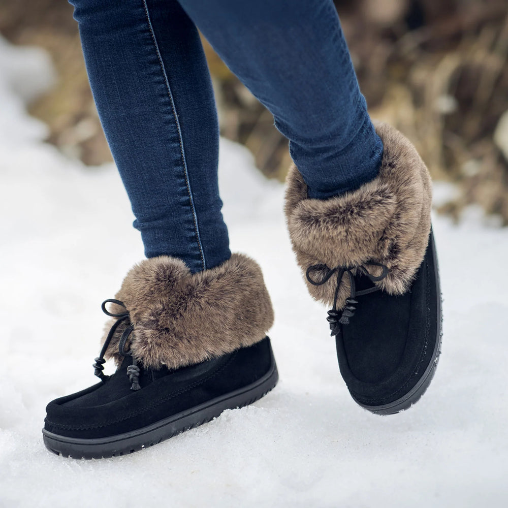 Person wearing black suede boots with brown fur trim and blue jeans, standing in snow.