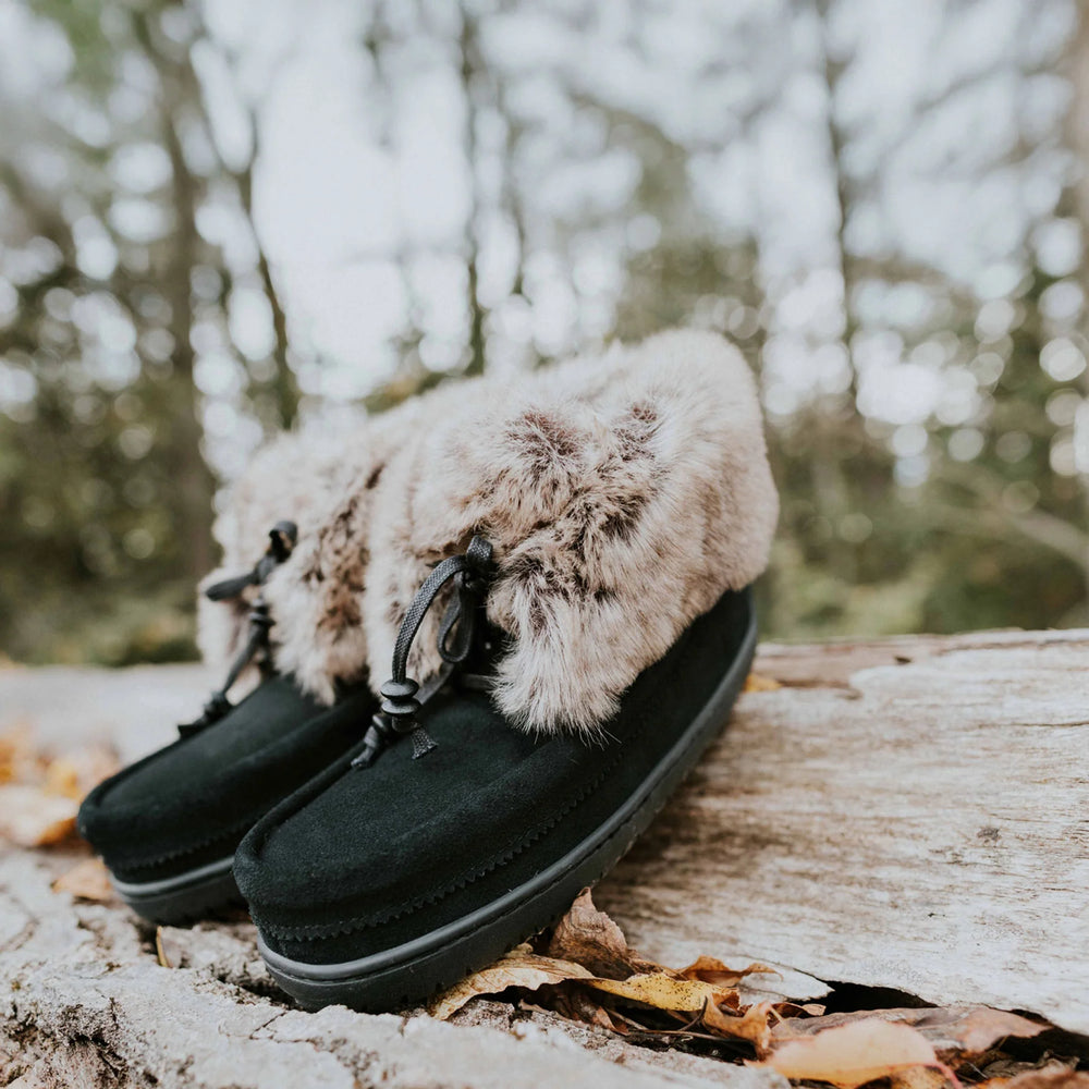 Black mukluk boots with faux fur trim rest on a fallen log surrounded by autumn leaves