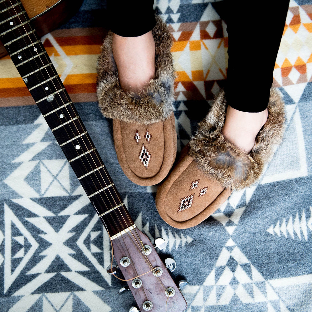 Fuzzy moccasins with fur trim and embroidered designs rest on a geometric patterned blanket next to an acoustic guitar neck, conveying cozy creativity.