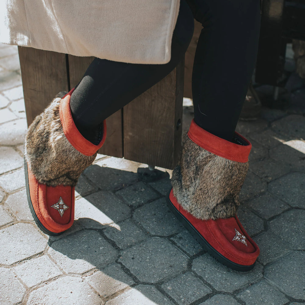 A person in black leggings and a beige coat sits on a wooden bench, wearing cozy red boots with fur trim and star details. The setting is sunny and inviting.