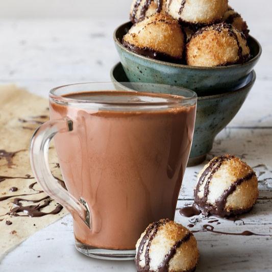 A ceramic mug filled with hot chocolate next to a bowl of golden-brown coconut macaroons drizzled with chocolate. Cozy and indulgent setting.