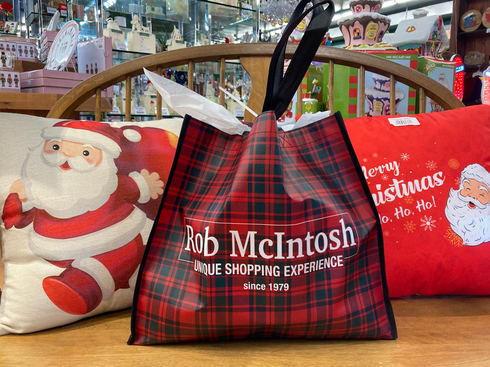 Red plaid shopping bag with 'Rob McIntosh' branding on a wooden surface with Christmas-themed pillows.