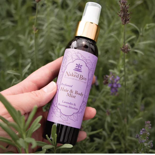 Hand holding a bottle of 'The Naked Bee' Perfumed Hair & Body Mist against a lavender background