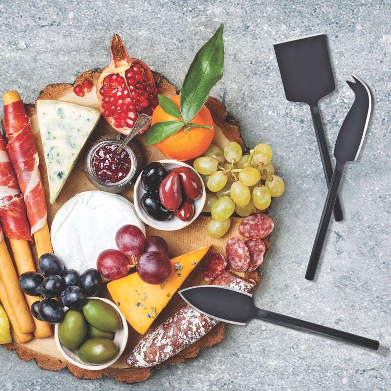 Assorted meats, cheeses, fruits, and olives on a wooden board with black cheese knives.