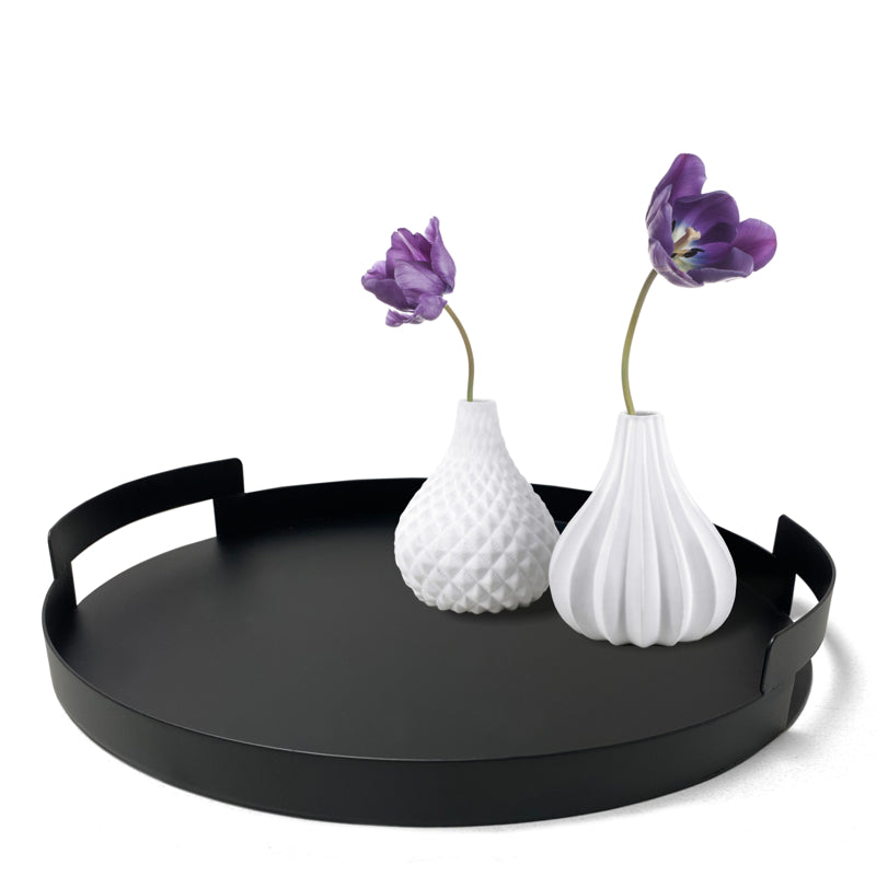 Black round tray with handles holding two white vases with purple flowers on a white background