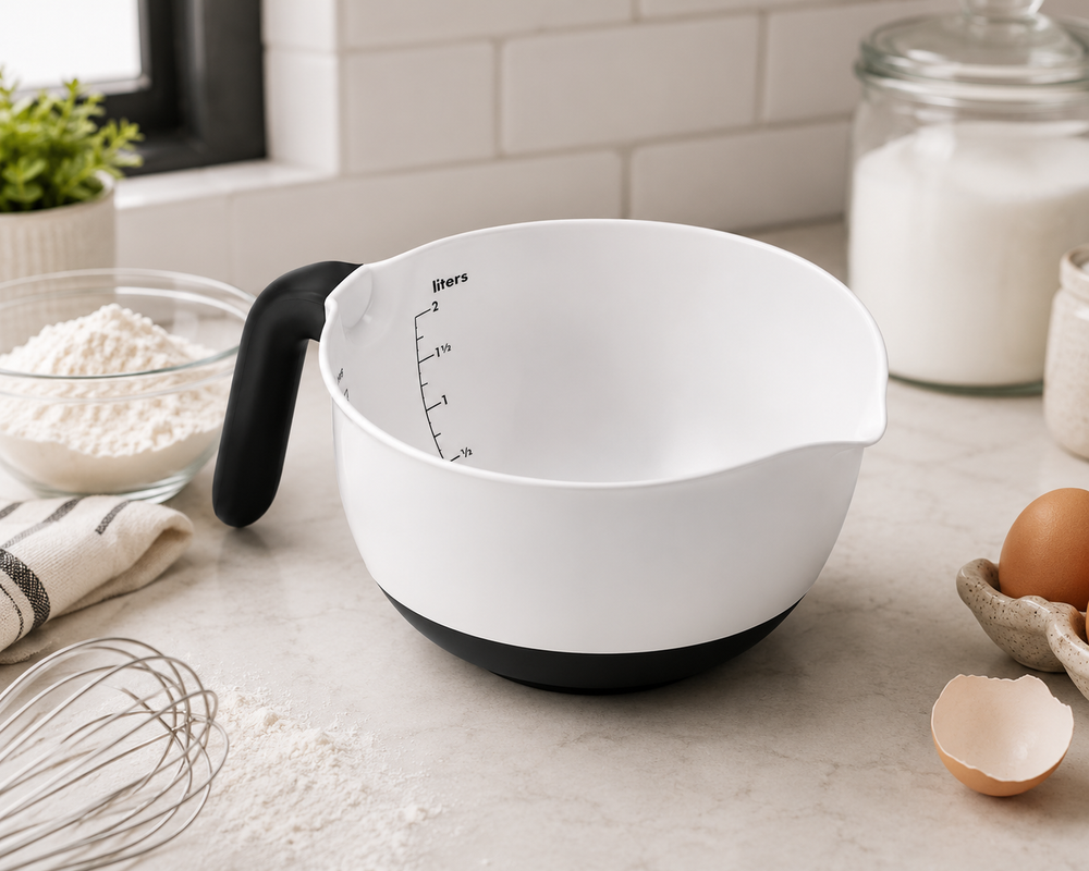 A white measuring bowl with a black handle and black base sits on a light stone countertop in a kitchen setting, surrounded by baking ingredients and utensils.