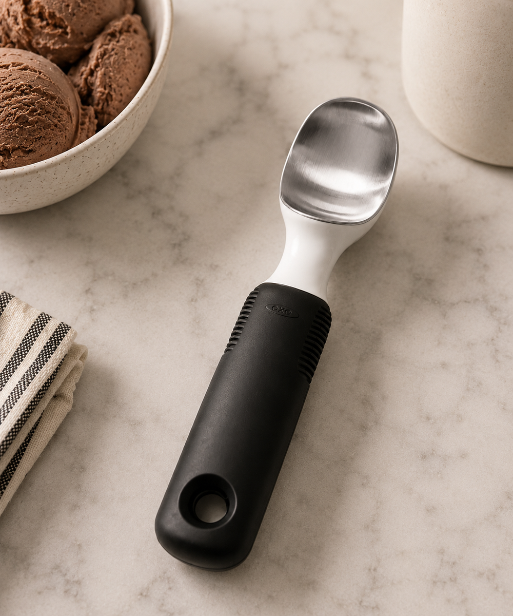 A kitchen ice cream scoop with a matte black handle and a polished stainless steel bowl, placed on a light marble surface with a bowl of ice cream and a striped napkin nearby.