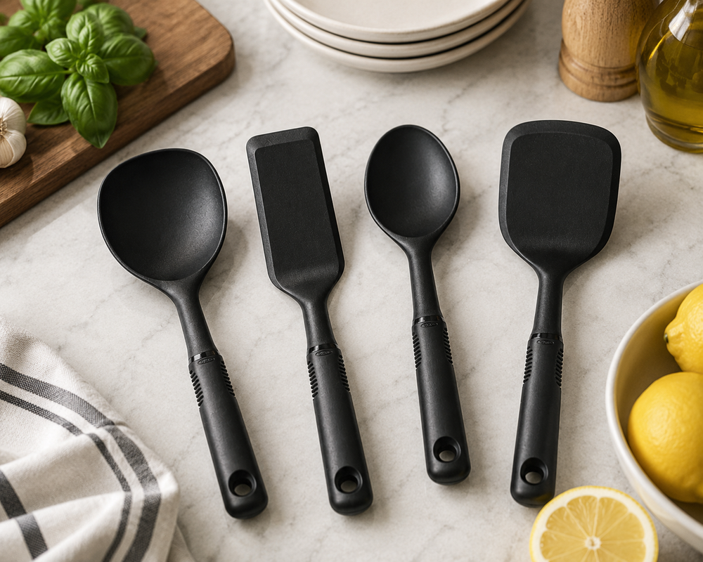 Four matte black silicone kitchen utensils, including two spoons and two scrapers, arranged on a light marble countertop with cooking ingredients and dishes around them.