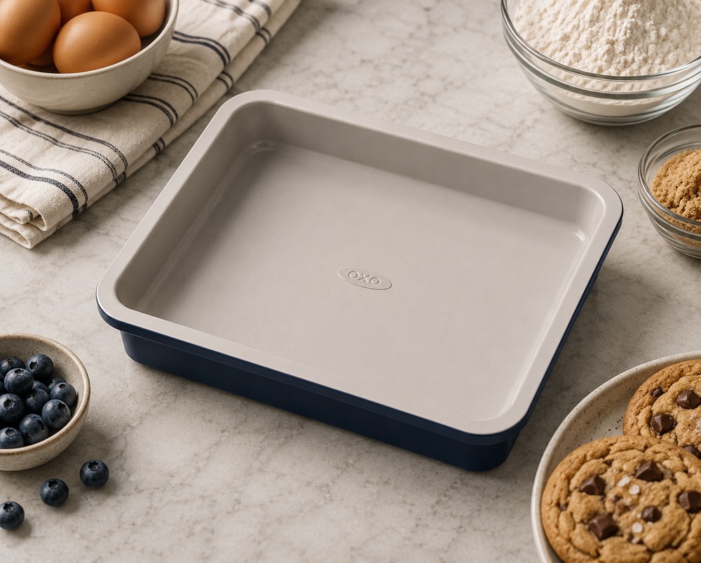 A square ceramic baking pan with a light gray interior and a navy blue exterior, styled on a marble countertop with baking ingredients and cookies around it.