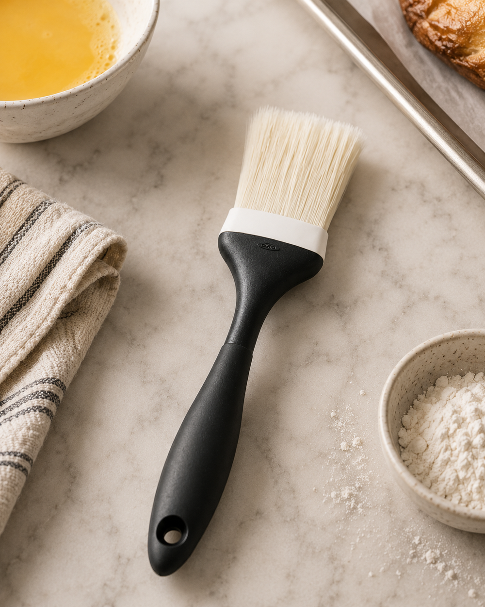 A silicone pastry brush with a matte black handle and white bristles, shown on a light marble surface with baking ingredients and kitchen props around it.