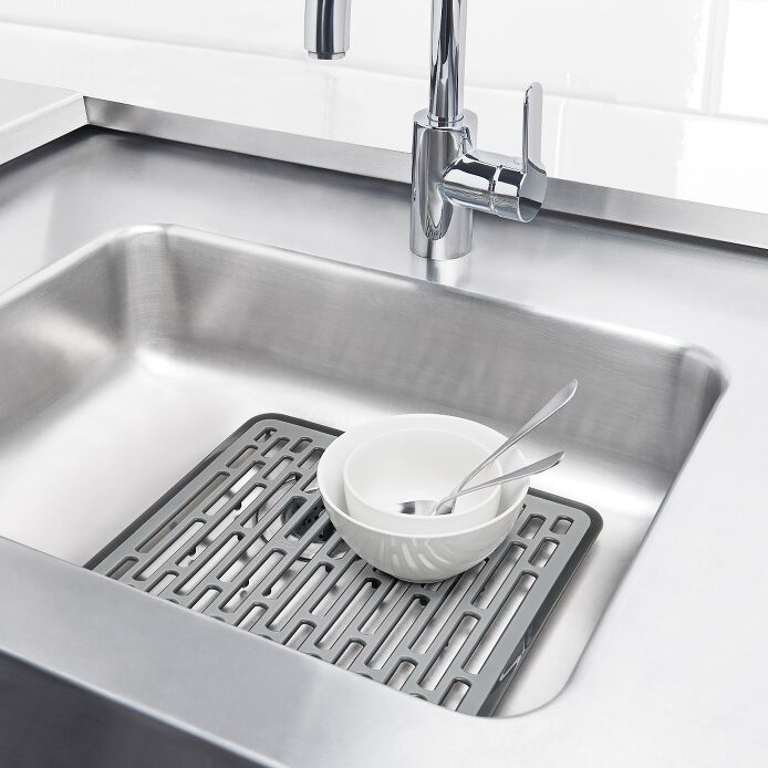 Stainless steel kitchen sink with a drying mat and bowl on a white tiled wall background
