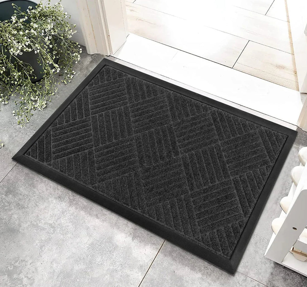 A black rectangular door mat with a black or grey pattern placed on a light floor at the entrance of a home.