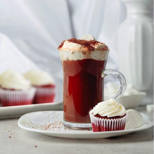 A glass mug of hot chocolate topped with cream and cocoa sprinkles sits on a plate with a red velvet cupcake and marshmallow.