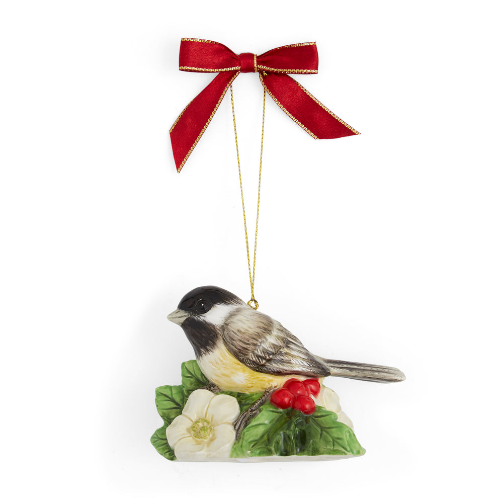 Porcelain ornament of a chickadee perched on green leaves with white flowers and red berries, suspended by a gold string and red bow. Festive and delicate.