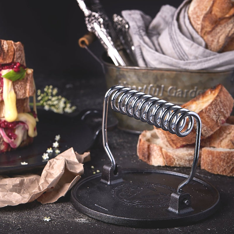 Cast iron bread press on a dark surface with bread and a sandwich in the background.