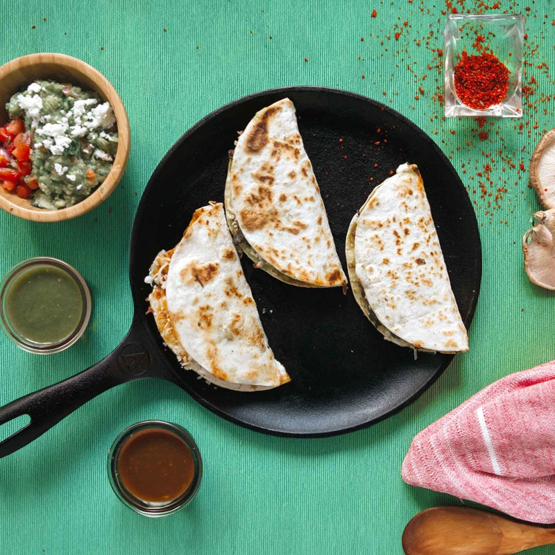 Three quesadillas on a black plate with various condiments on a green surface