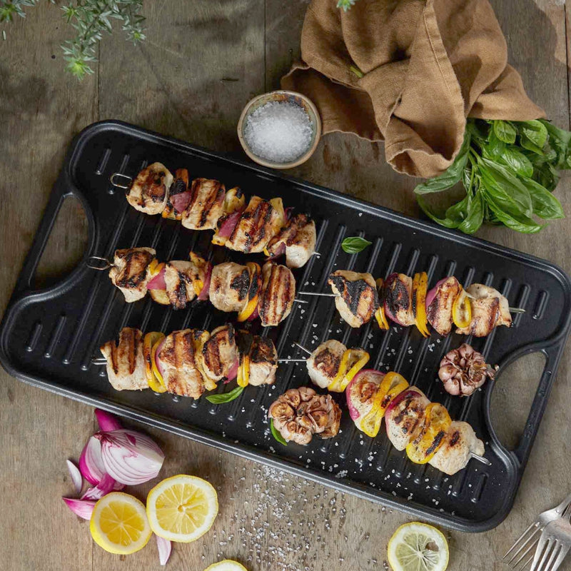 Grilled skewers on a black grill pan with lemon slices and herbs on a wooden surface.