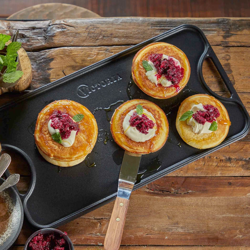 Four small pancakes with toppings on a black tray with 'Victoria' branding, on a wooden surface.