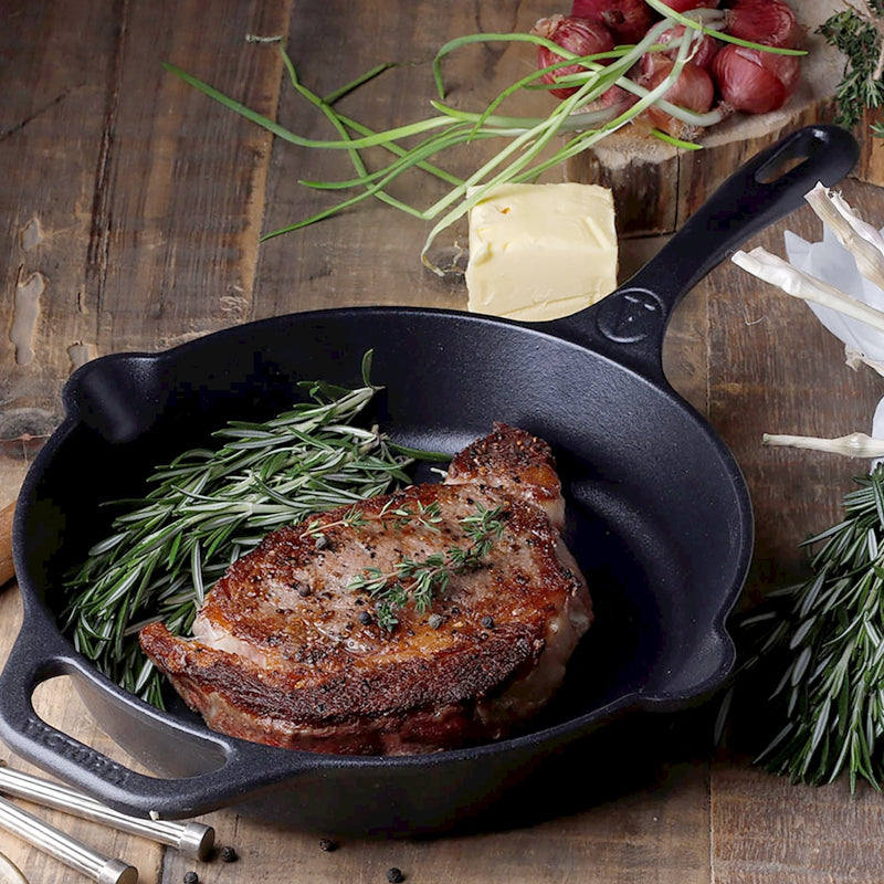 Grilled meat with rosemary in a black cast iron skillet on a wooden surface.